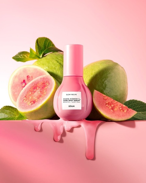 A high-end product photography shot featuring a dark spot serum bottle surrounded by sliced guavas with a liquid serum spill on a pink surface.